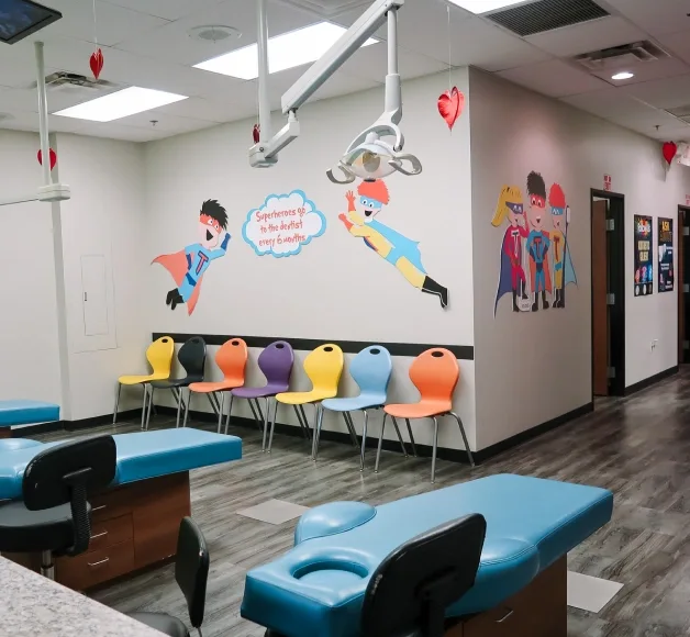 Pediatric dental treatment area featuring blue exam tables, colorful wall art with superhero characters, and vibrant seating.
